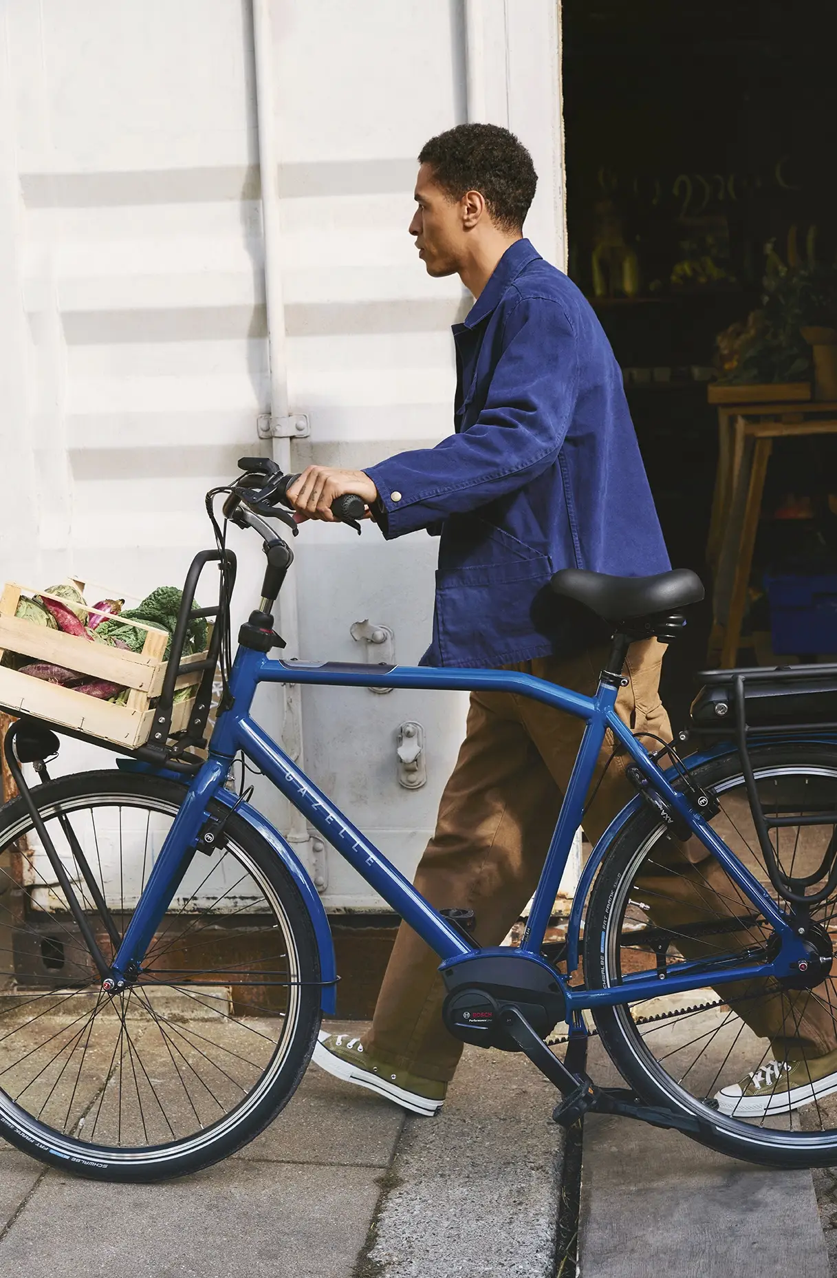 A person in a blue jacket walks alongside a blue bicycle with a front crate filled with produce, including broccoli and other greens.