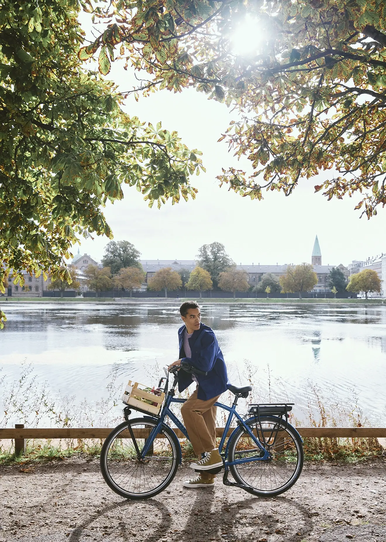 Man on a bicycle pauses by a scenic lake under sunlit trees, with a city skyline and blue sky in the background.
