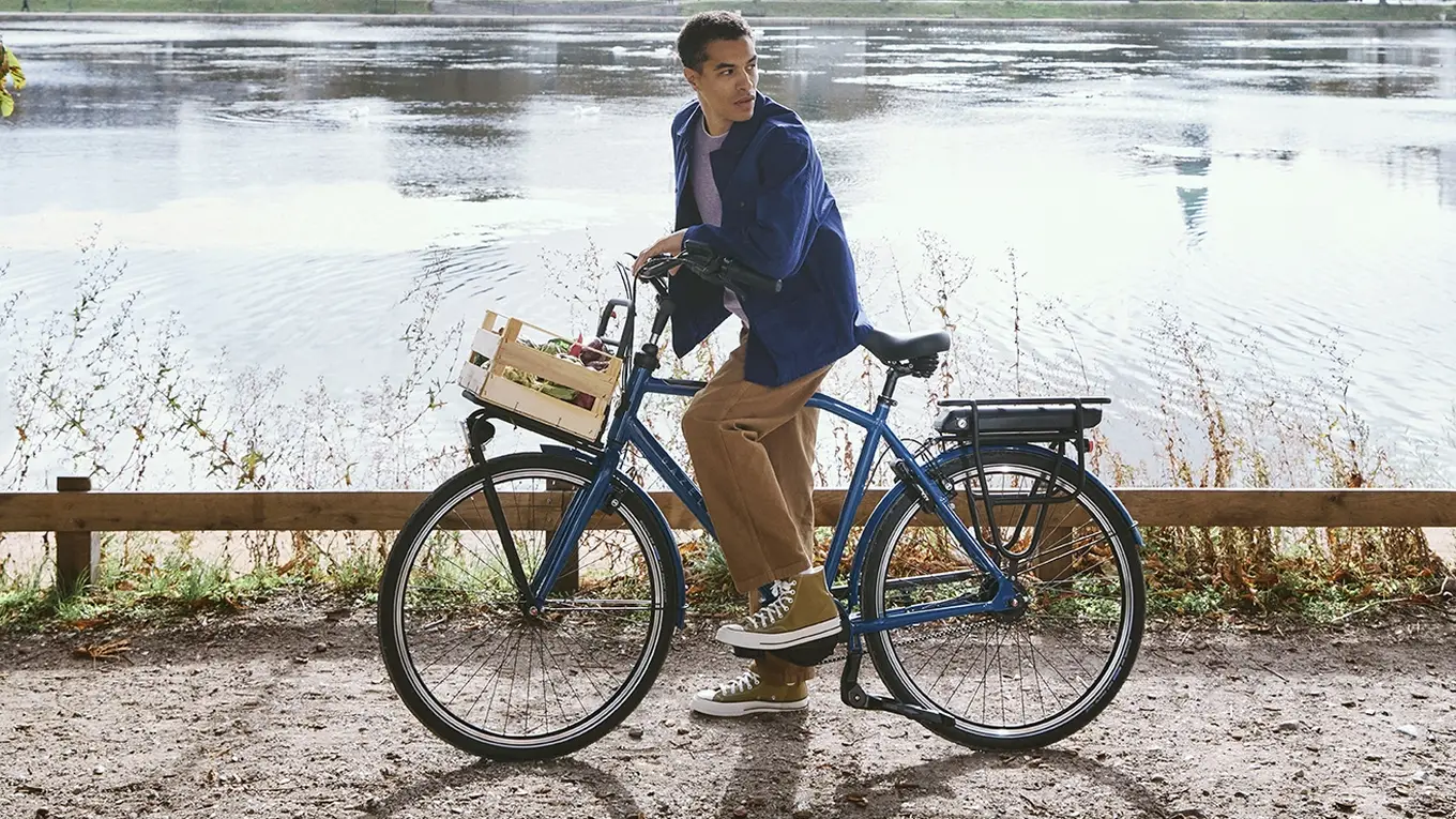Man on a bicycle pauses by a scenic lake under sunlit trees, with a city skyline and blue sky in the background.
