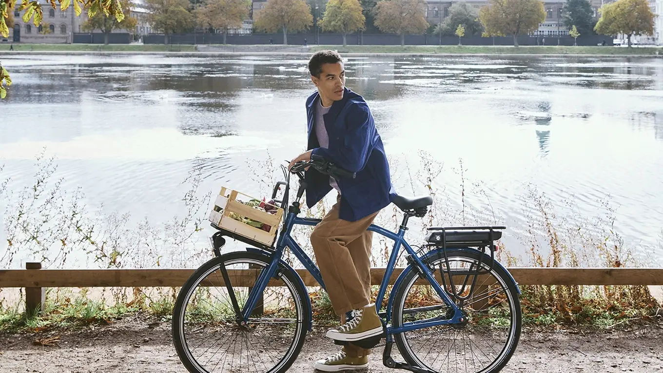 Man on a bicycle pauses by a scenic lake under sunlit trees, with a city skyline and blue sky in the background.
