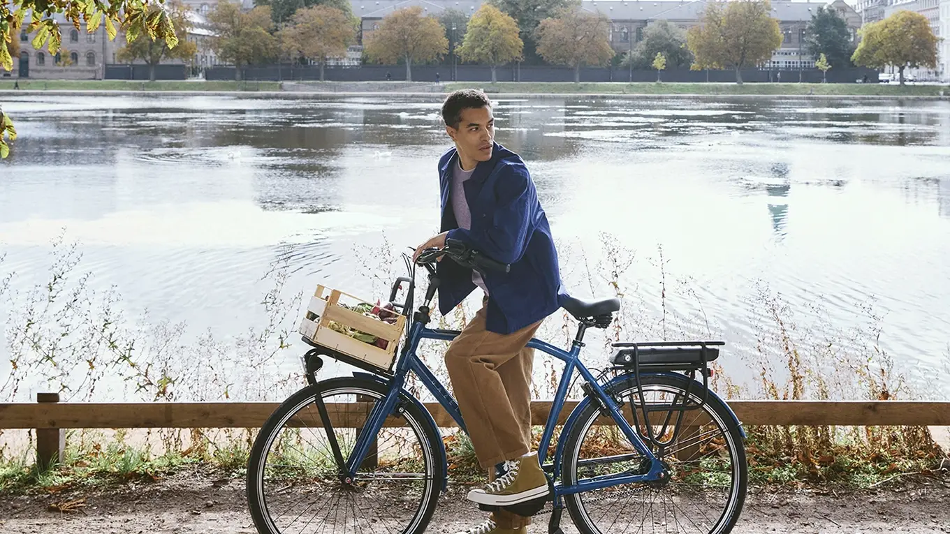 Man on a bicycle pauses by a scenic lake under sunlit trees, with a city skyline and blue sky in the background.