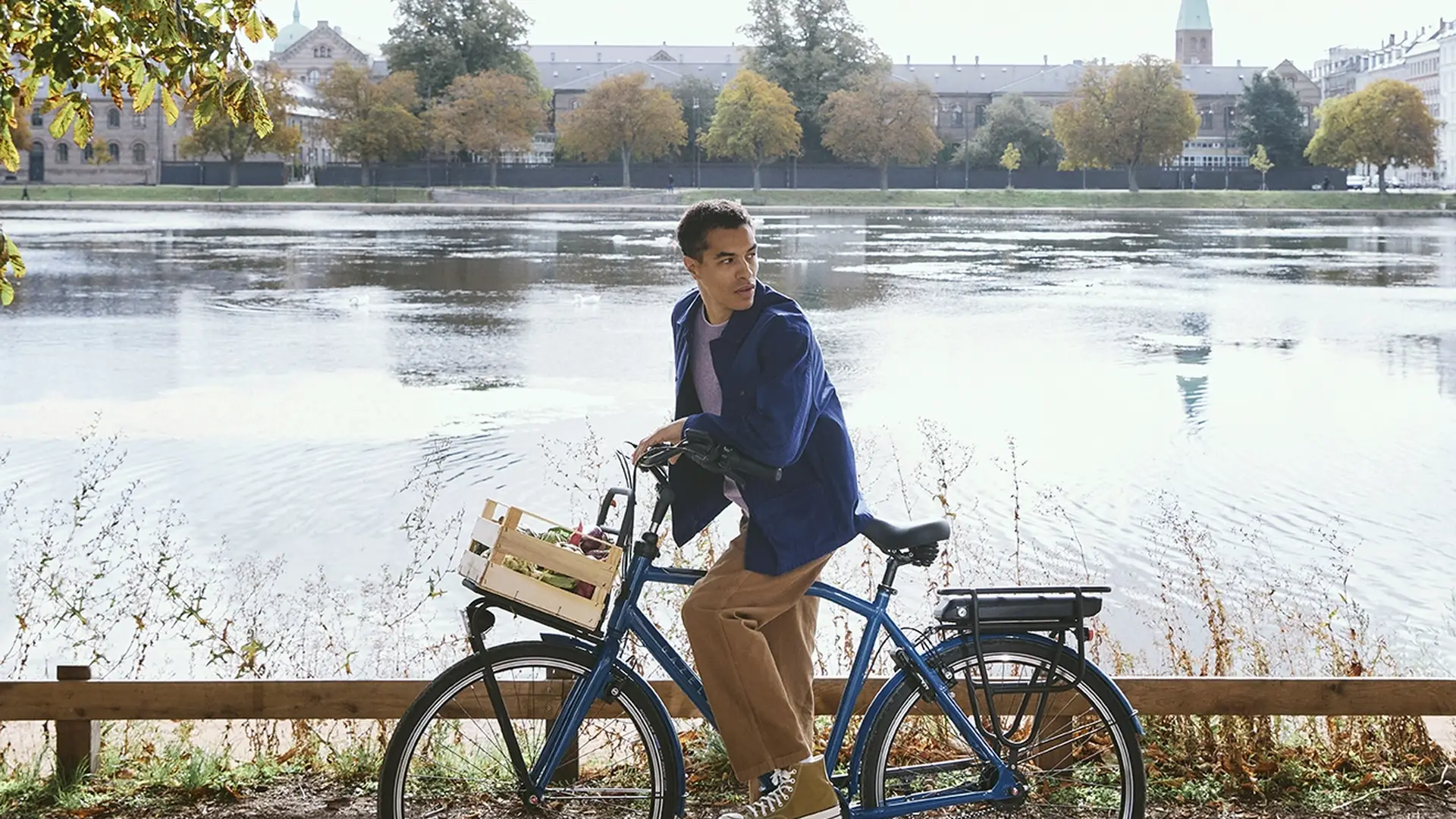 Man on a bicycle pauses by a scenic lake under sunlit trees, with a city skyline and blue sky in the background.