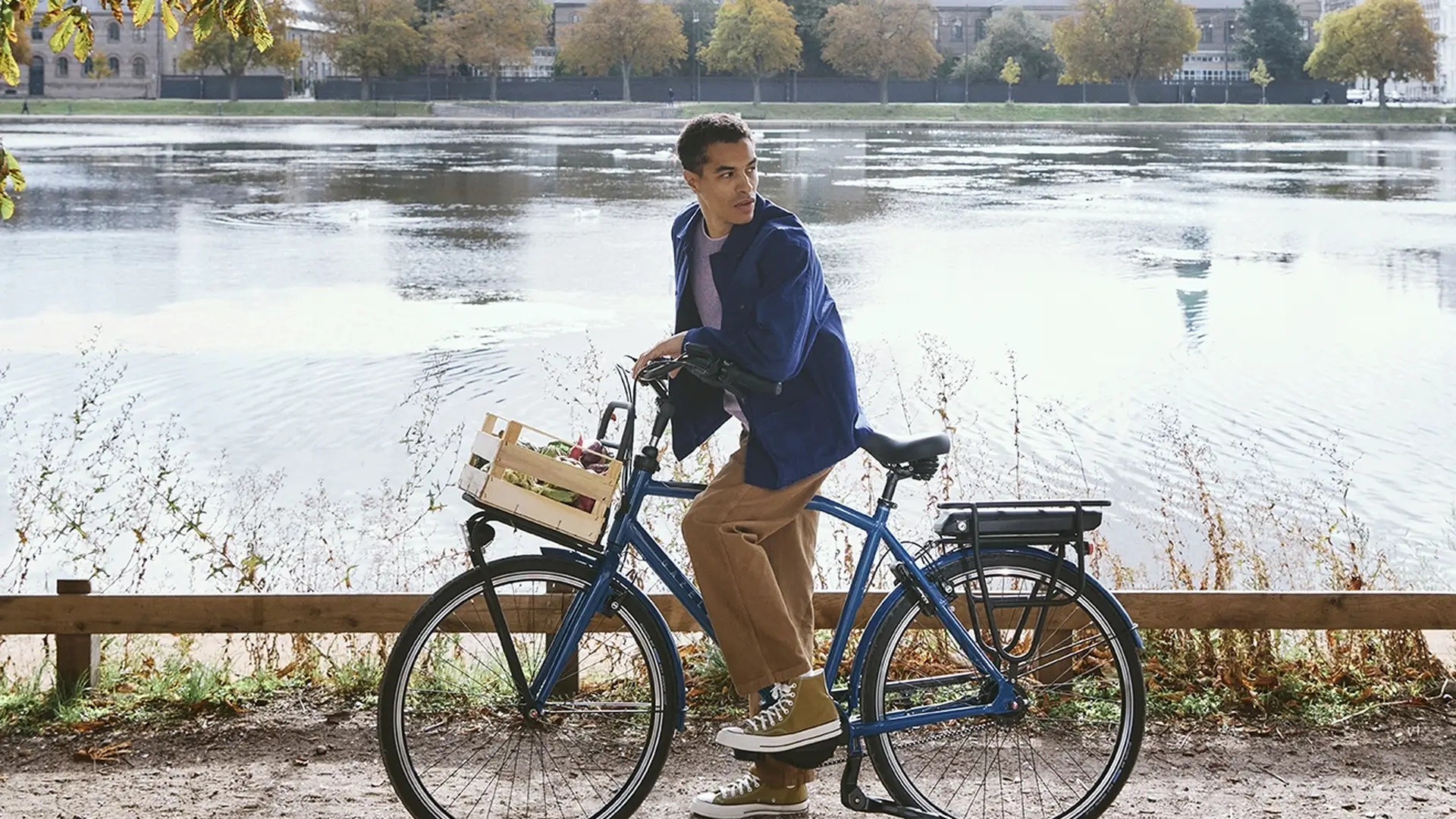 Man on a bicycle pauses by a scenic lake under sunlit trees, with a city skyline and blue sky in the background.