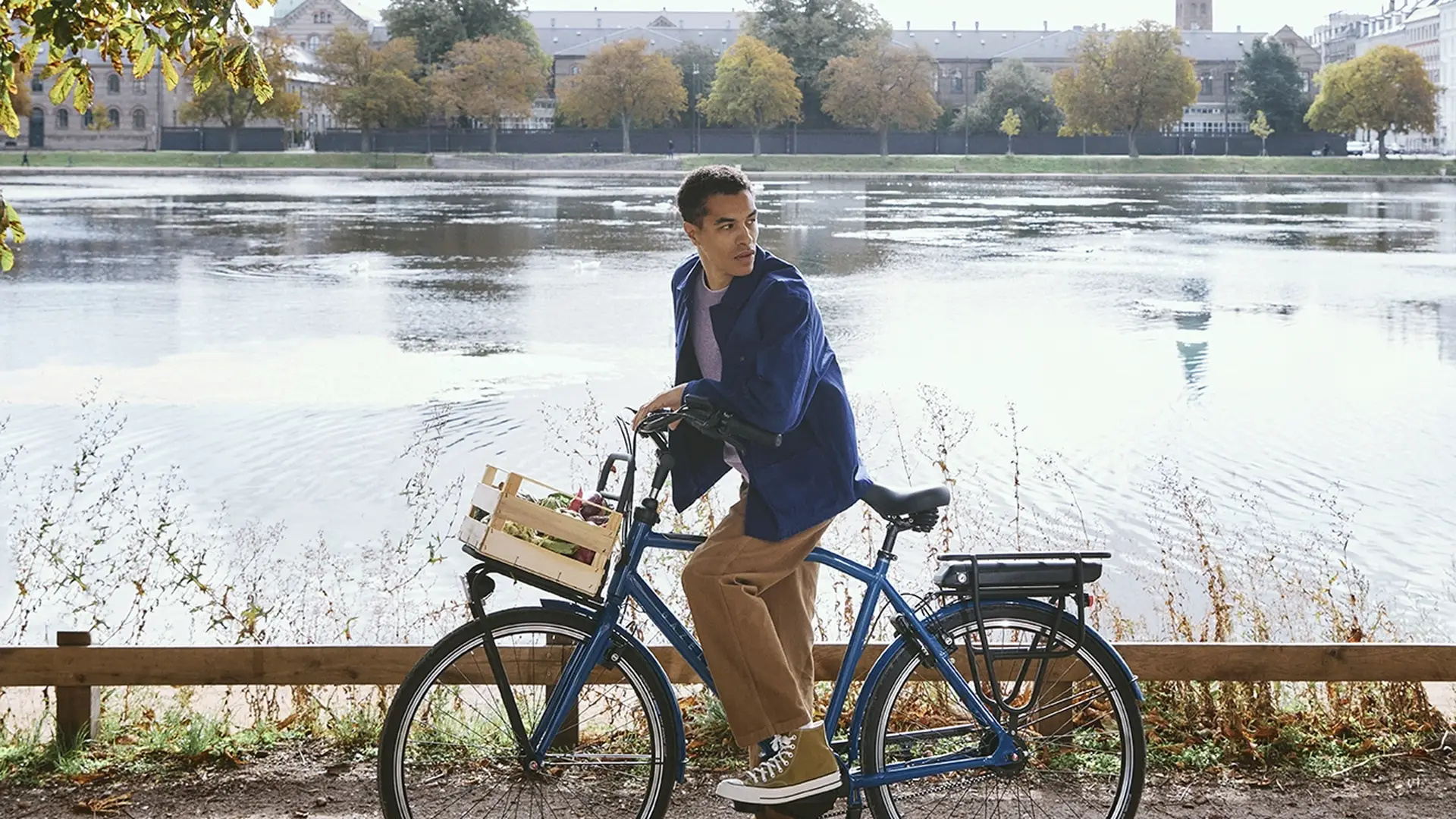 Man on a bicycle pauses by a scenic lake under sunlit trees, with a city skyline and blue sky in the background.