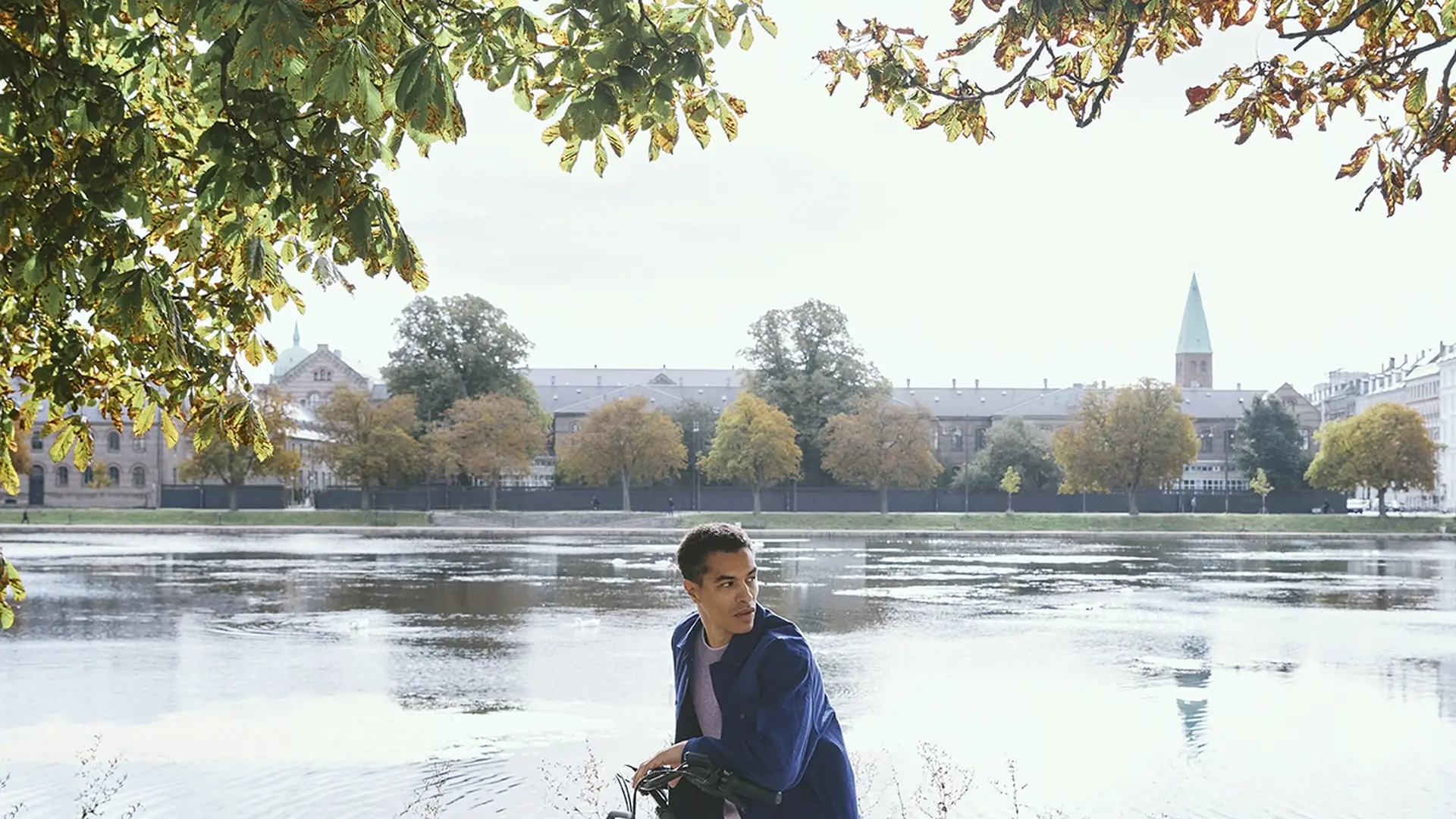 Man on a bicycle pauses by a scenic lake under sunlit trees, with a city skyline and blue sky in the background.