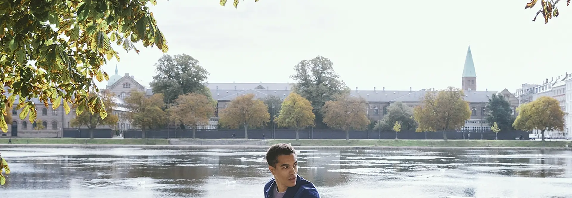 Man on a bicycle pauses by a scenic lake under sunlit trees, with a city skyline and blue sky in the background.