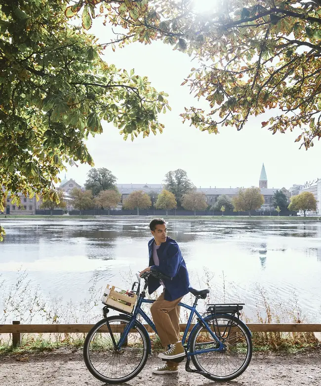 Man on a bicycle pauses by a scenic lake under sunlit trees, with a city skyline and blue sky in the background.