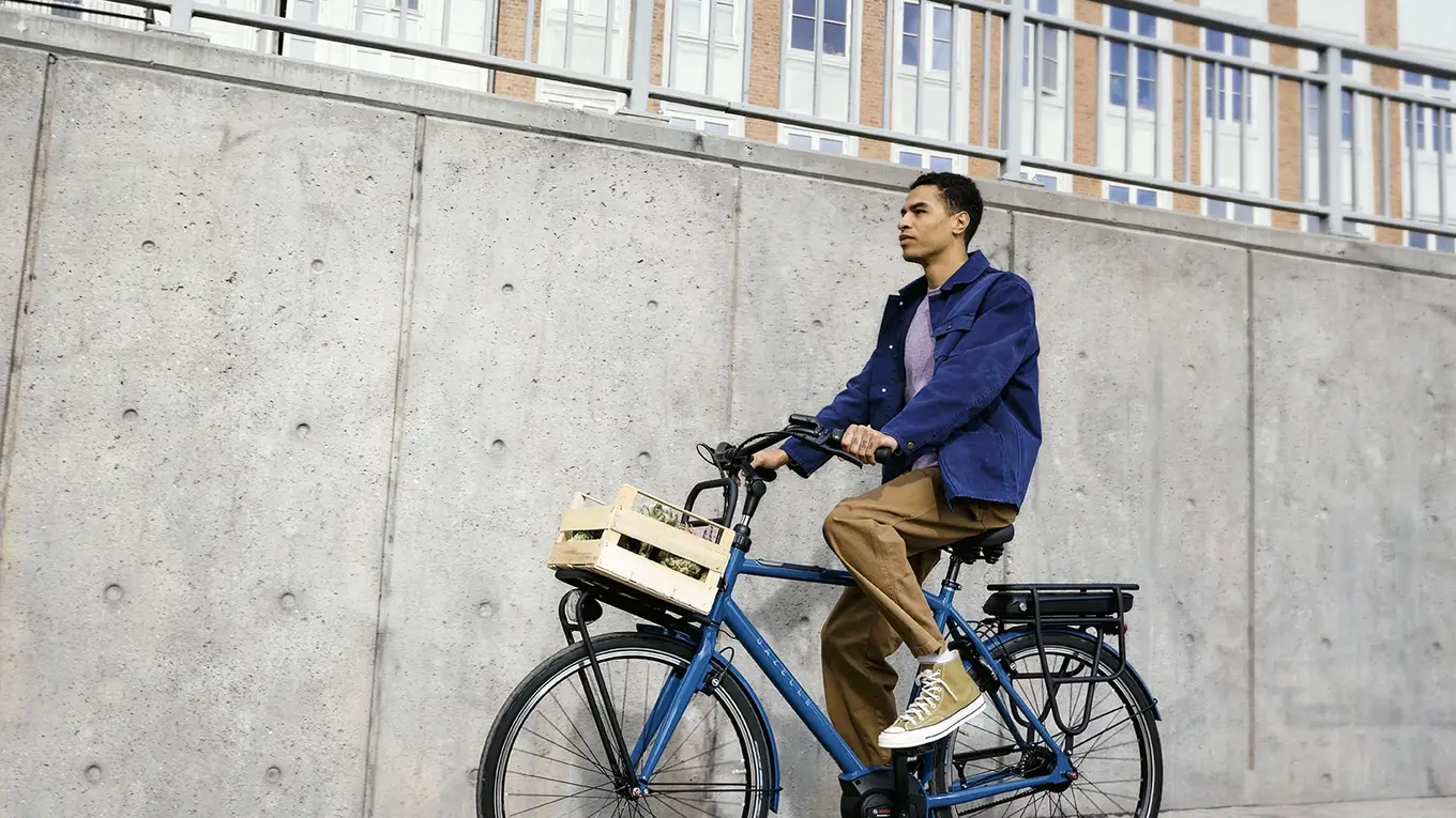 Person riding a blue bicycle with a wooden basket in an urban area, wearing a blue jacket and brown pants, near a concrete wall.