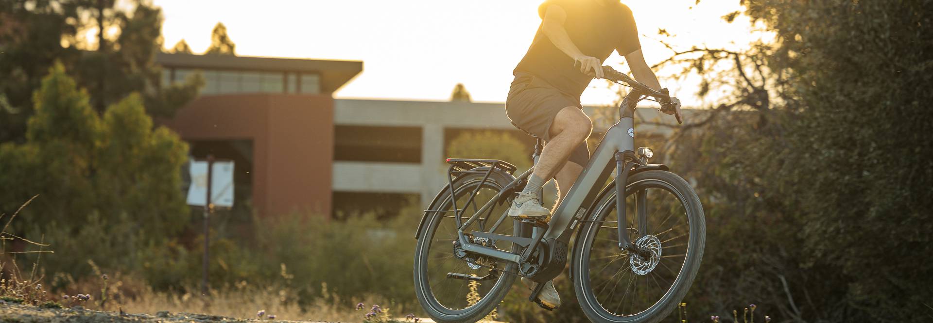 Person riding an electric bike on a sunny trail, surrounded by trees and a modern building in the background.