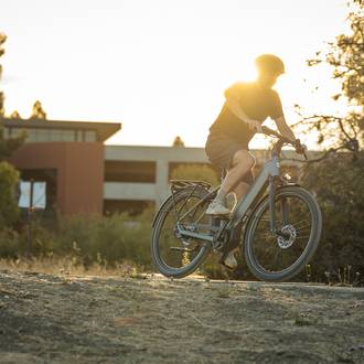 Person riding an electric bike on a sunny trail, surrounded by trees and a modern building in the background.