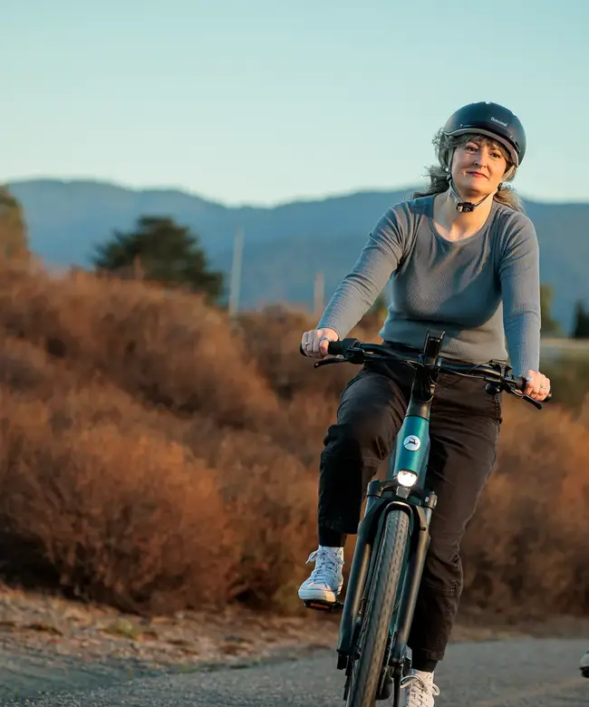 Two people wearing helmets are riding bicycles on a path bordered by dry bushes and distant mountains under a clear sky.