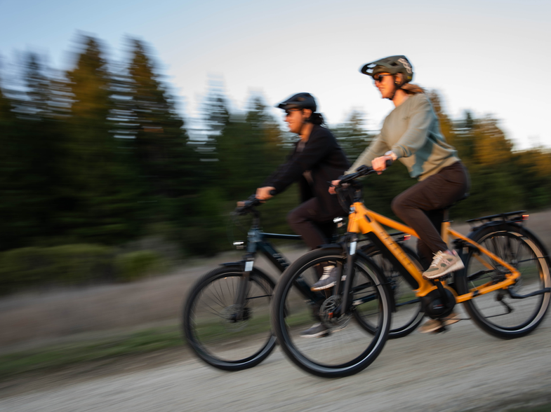 Two people cycling on a gravel path through a forest, wearing helmets and casual clothing, with motion blur indicating speed.