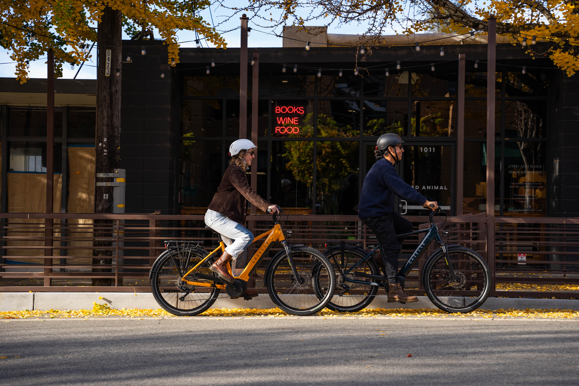 Two people ride bicycles on a street lined with autumn trees. A building with a sign reading "Books Wine Food" is in the background.