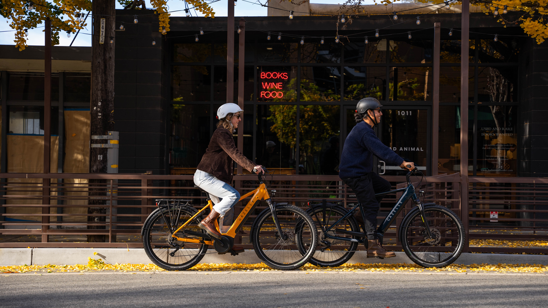 Two people ride bicycles on a street lined with autumn trees. A building with a sign reading "Books Wine Food" is in the background.
