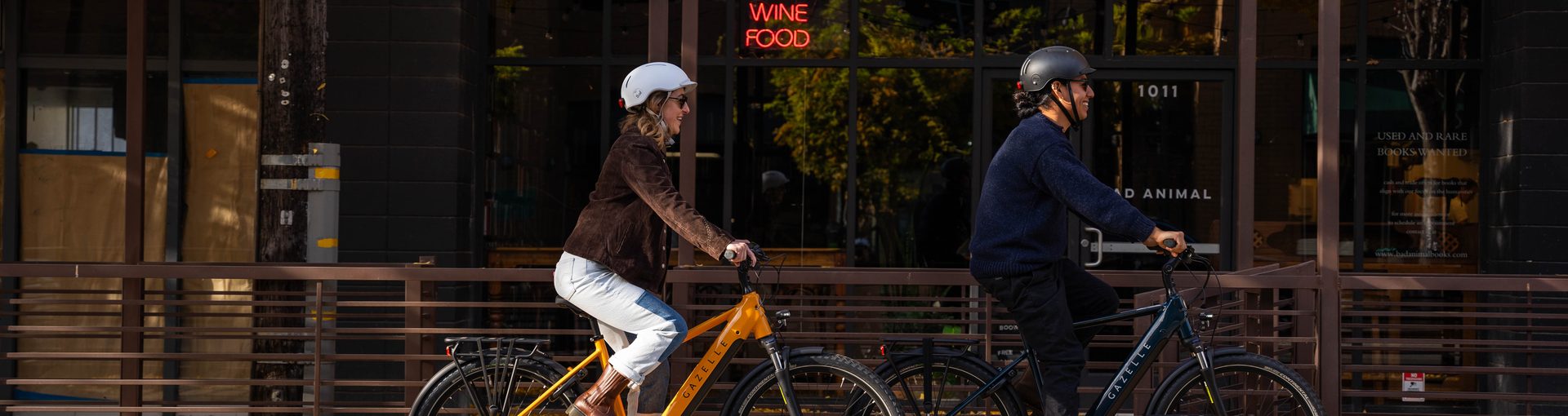 Two people ride bicycles on a street lined with autumn trees. A building with a sign reading "Books Wine Food" is in the background.