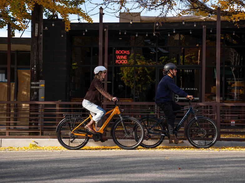 Two people ride bicycles on a street lined with autumn trees. A building with a sign reading "Books Wine Food" is in the background.