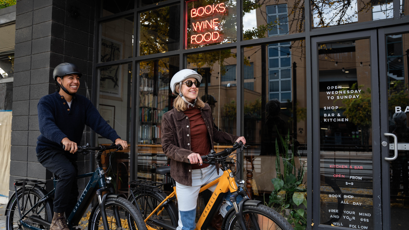 Two people on bicycles smile in front of a shop with a neon sign reading "Books, Wine, Food." The storefront has large windows and leafy decor.