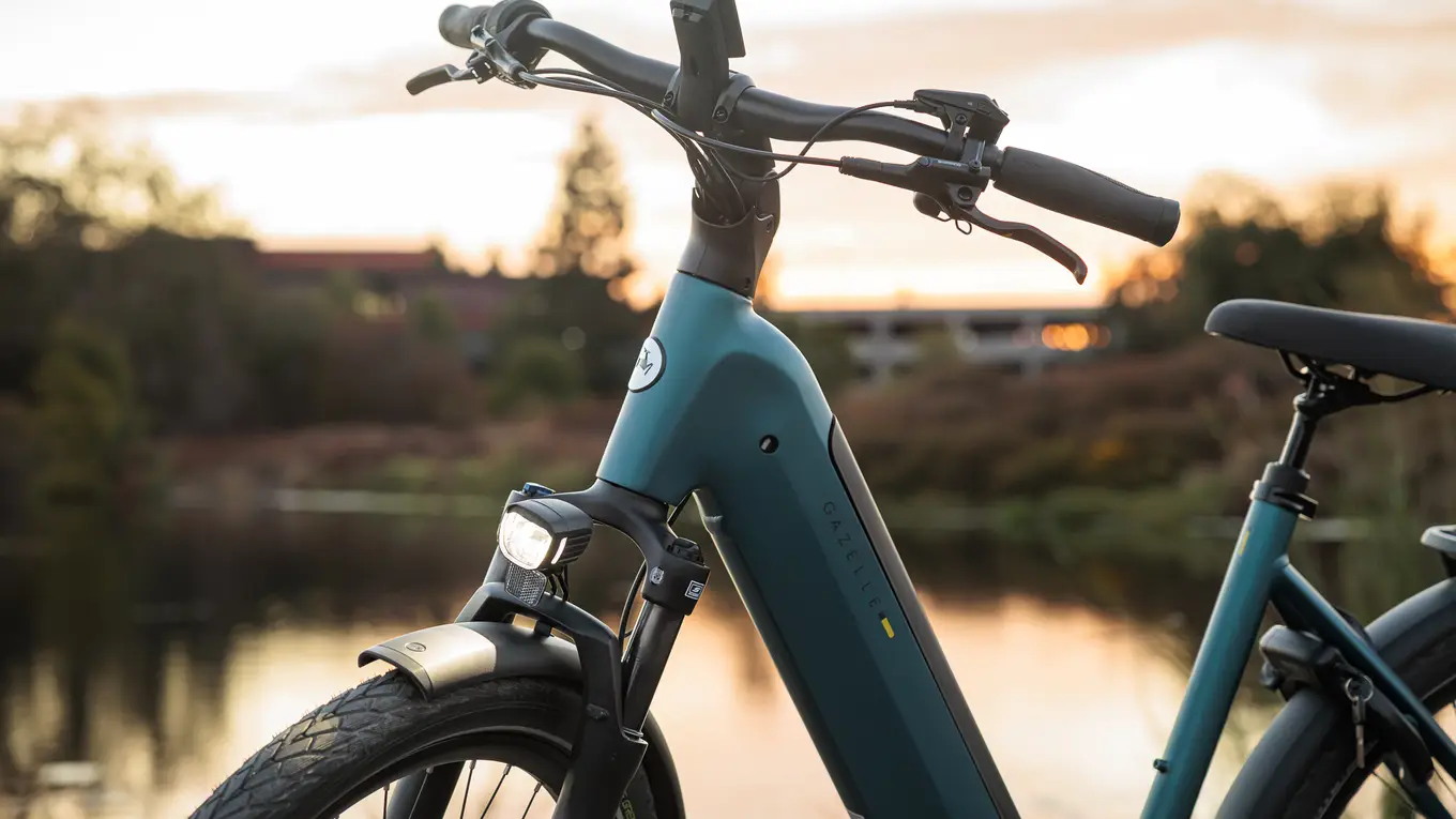 Close-up of a teal electric bicycle near a lake at sunset, highlighting the handlebars and front light, with blurred trees in the background.